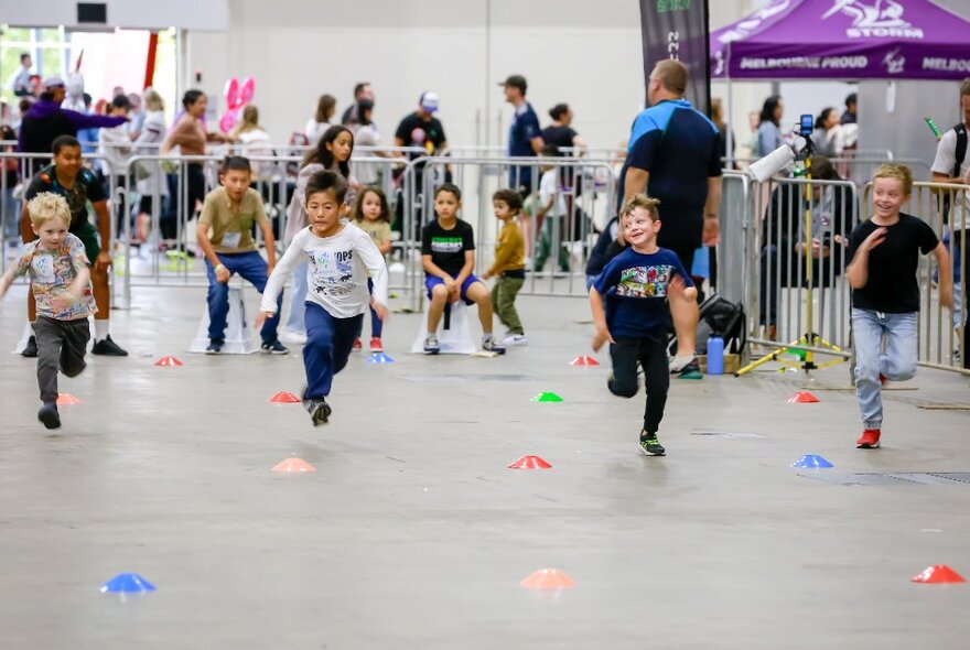 Children in a running race in a large venue with barricades and onlookers.