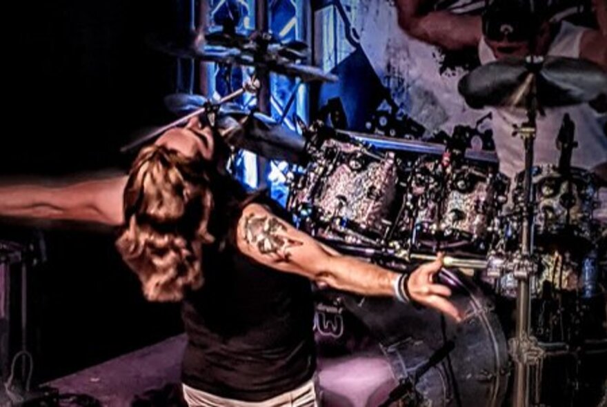 A singer performing on stage, leaning back into the audience with outstretched arms and a drummer in the background.