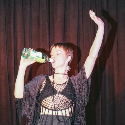 A performer on stage in front of a red curtain, drinking from an open wine bottle, one arm raised above her head.