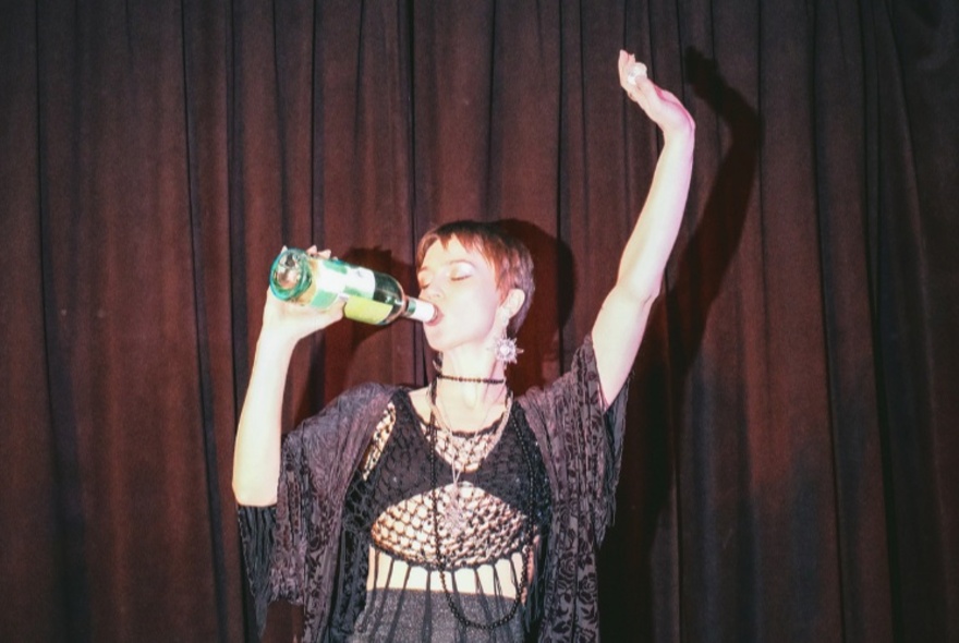 A performer on stage in front of a red curtain, drinking from an open wine bottle, one arm raised above her head.