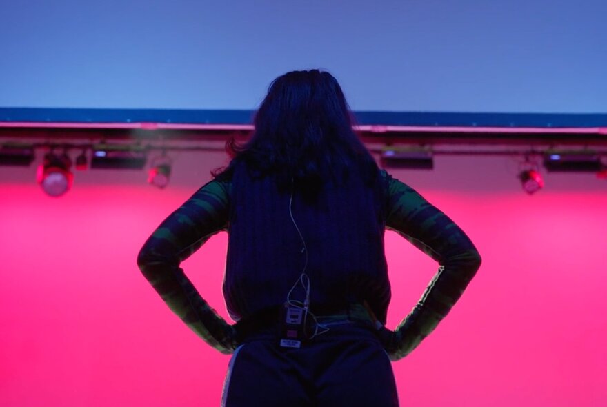 A silhouette of a person with long dark hair, seen from behind, standing on a stage against a vibrant pink background.