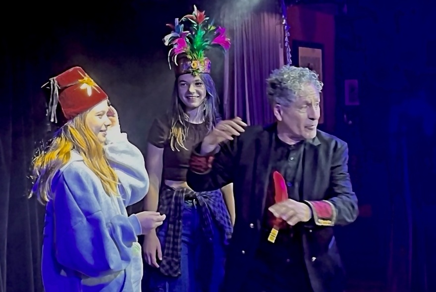 A man performing a magic show with two teenage girls assisting.