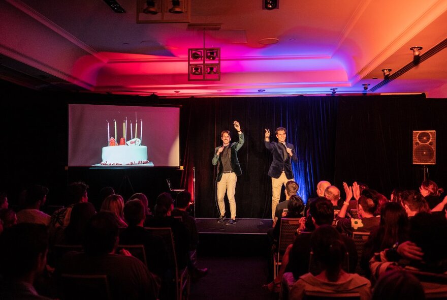 Two male comedians dressed the same, on stage, with a slide presentation of a birthday cake with candles behind them.