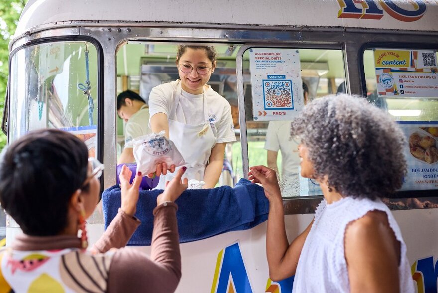 People buying produce from a woman through the window of a cream-coloured converted bus.