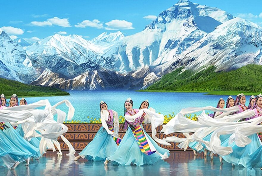 Classical Chinese dancers in long blue dresses all performing on a stage with a set depicting snow covered mountains and a lake. 