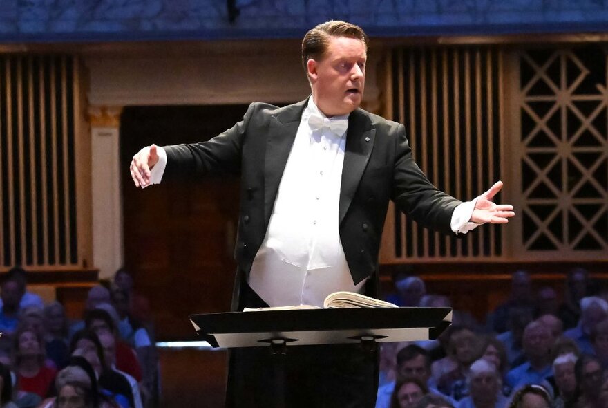 An orchestra conductor wearing a tuxedo, using his hands to conduct, an audience seated behind him.