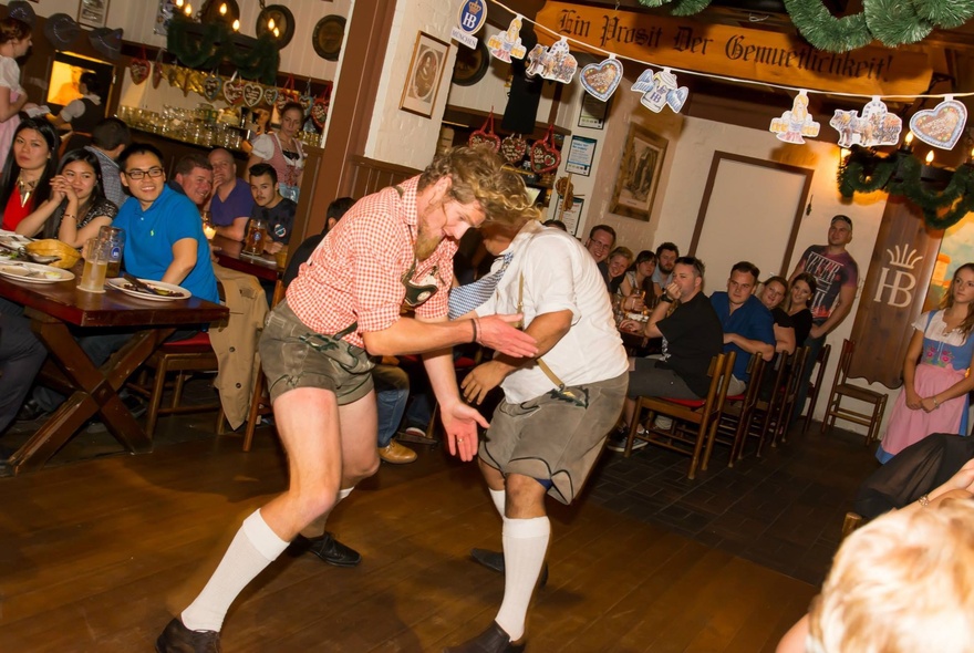 Two men wearing Lederhosen wrestle dancing while customers look on.