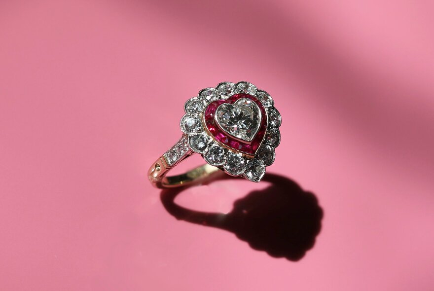 A diamond ring in the shape of a heart with a pink stone inlay, against a pink background with a shadow cast. 