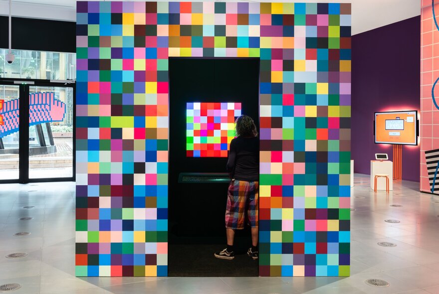 A person wearing multi-coloured shorts watching a screen with a multi-colour pixellated pattern in a booth decorated with the same pattern.