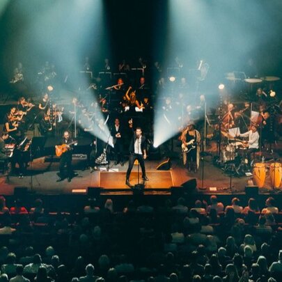 A live performance of The Cat Empire band alongside a full symphony orchestra on a large stage, in front of a seated audience.