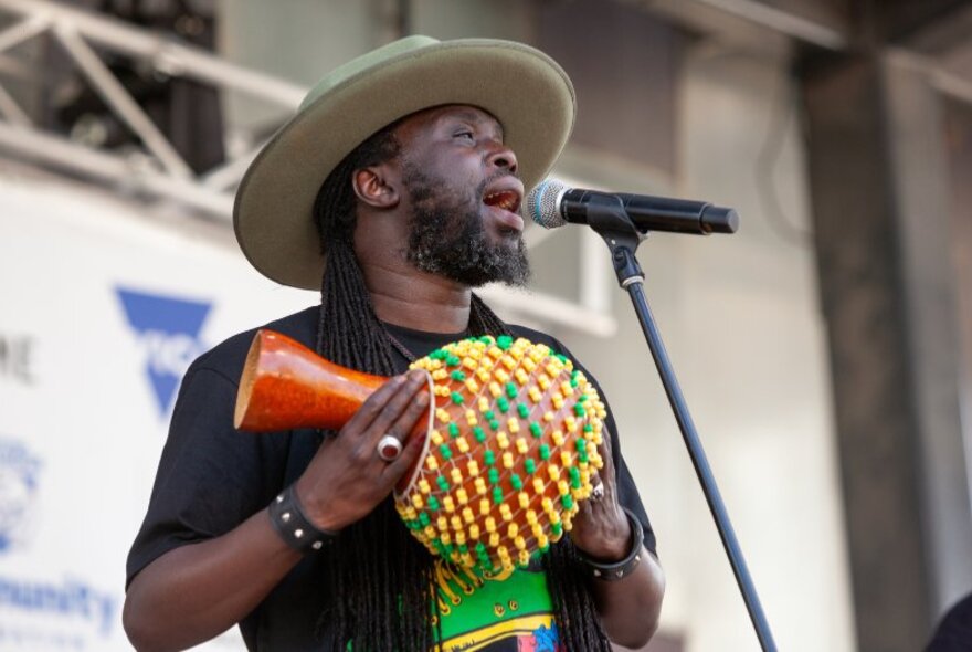 A man with a hat singing on stage and playing a traditional African instrument. 
