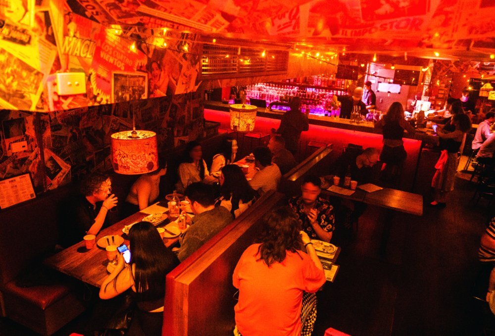 Underground Mexican restaurant with a dimly lit glowing red interior, funky wallpaper and lots of customers drinking and dining.