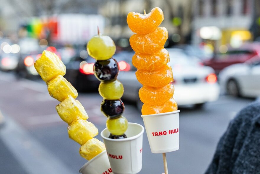 Candied pineapple, grapes and mandarin slices on sticks in paper cups.