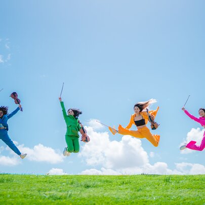 The four members of Invictus Quartet, each wearing a different coloured suit, jumping up in the air from a grassy hill, while holding their instruments, against a blue sky background.