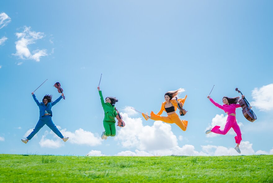 The four members of Invictus Quartet, each wearing a different coloured suit, jumping up in the air from a grassy hill, while holding their instruments, against a blue sky background.