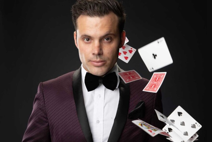 A magician against a black background with playing cards flying through the air.