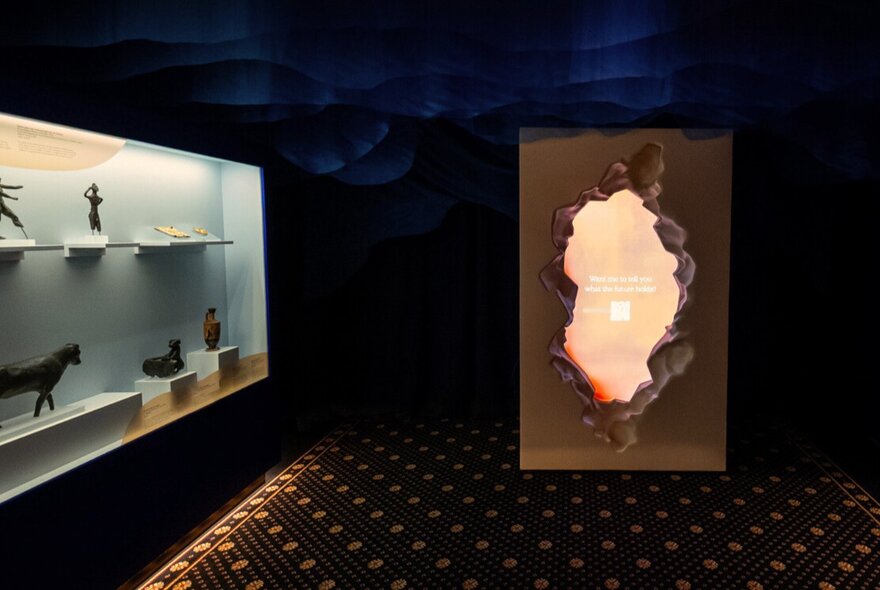 A room at the Hellenic Museum in Melbourne, with ancient artefacts in a glass cabinet on the left and an installation on the right that allows the viewer to look through a pretend hole in the wall.