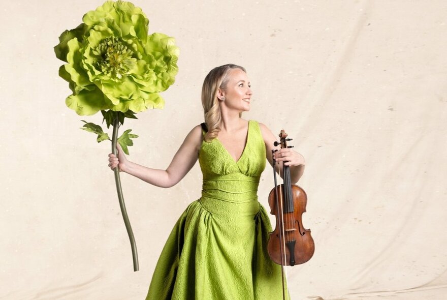 A woman in a lime green evening dress holds a violin in one hand and a giant lime green flower in the other. 