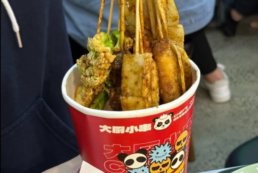 Fried and barbecued skewers in a takeaway paper cup.