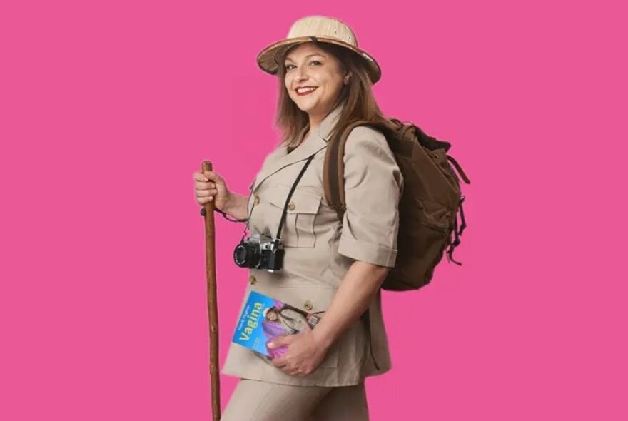 A woman dressed in a safari-style outfit, including a pith helmet, backpack, and camera, and carrying a travel guidebook in her hand, posed against a pink background.