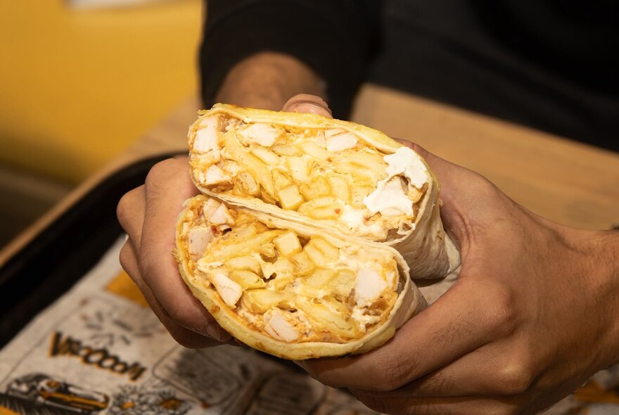 A person holding a cut chicken and French fries wrap or French taco over a table.