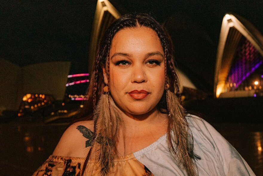 Singer Bumpy, standing outdoors at night, the Sydney Opera House just visible behind her, slightly smiling and looking directly at the viewer, her hair plaited on either side of her face.