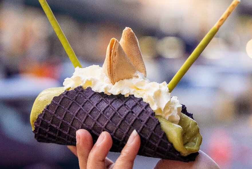 A hand holding a chocolate wafer like taco filled with ice cream and garnished with whipped cream and a fortune cookie.