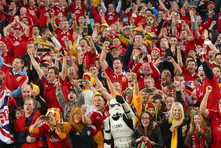 Rugby fans in a packed sports stadium, wearing their team colours of red, cheering and waving.