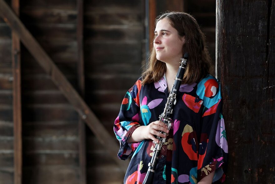 Woman wearing a dark dress with brightly coloured pattern, holding a clarinet in a wooden building.