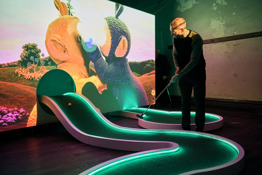A person playing on a curving mini-golf putting green, highlighted in green neon coming out of an arched hole in a projector screen that shows two Teletubbies in a field. 