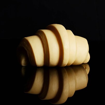 An unbaked croissant, on a reflective black surface in front of a black background.