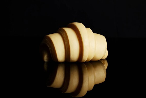An unbaked croissant, on a reflective black surface in front of a black background.