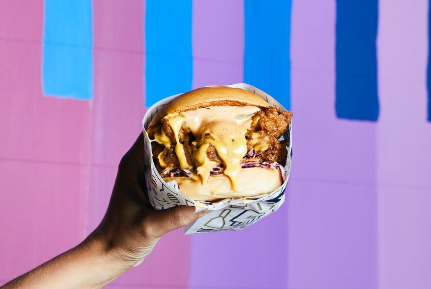 Hand holding a hamburger wrapped in paper, with melted cheese dripping out the sides.