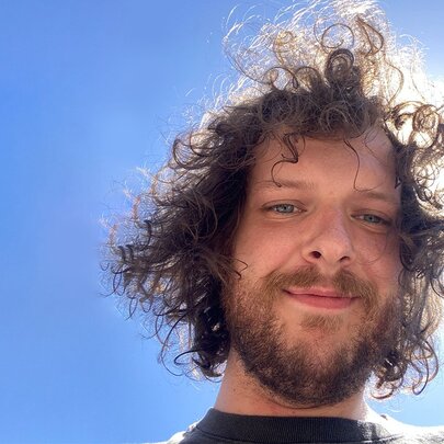 Portrait of comedian Blake Everett looking down at the camera, the sun behind his head in a bright blue sky.