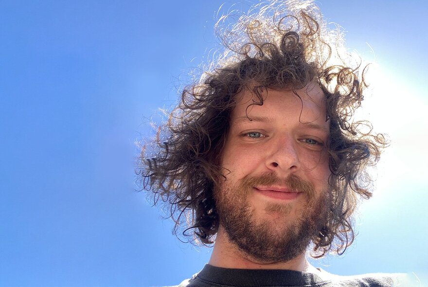 Portrait of comedian Blake Everett looking down at the camera, the sun behind his head in a bright blue sky.