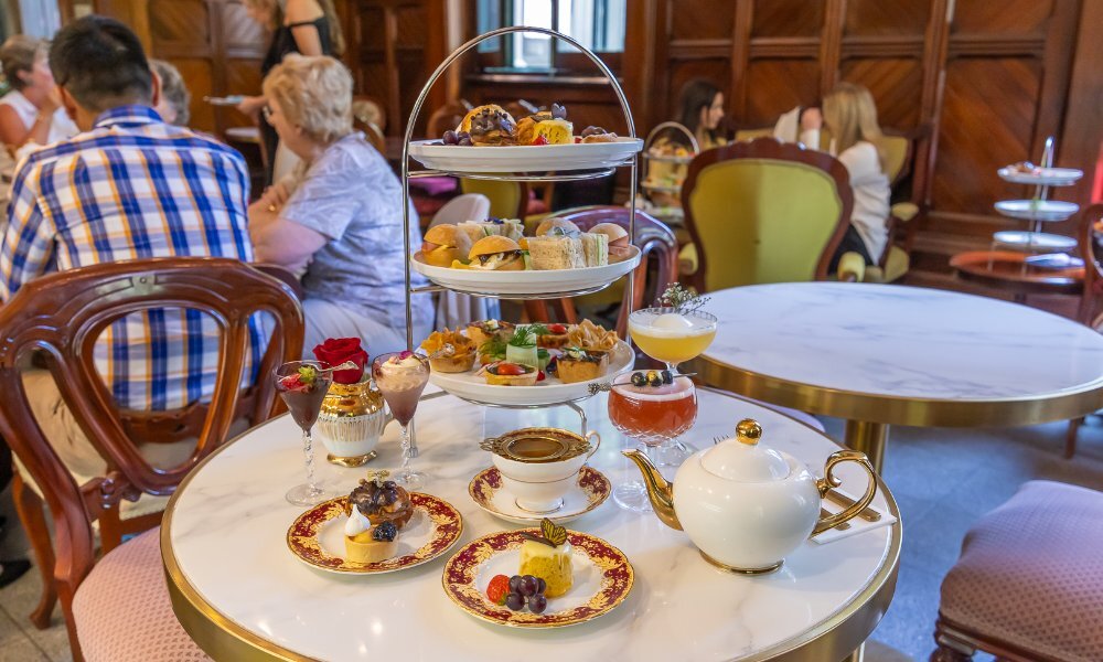 A white marble table in a restaurant with a three-tier cake stand holding small high tea snacks, and an assortment of cocktails, tea and desserts.