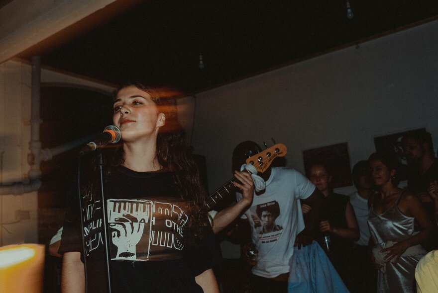 A woman singing into a microphone with someone behind her playing guitar, a small group of people watching.