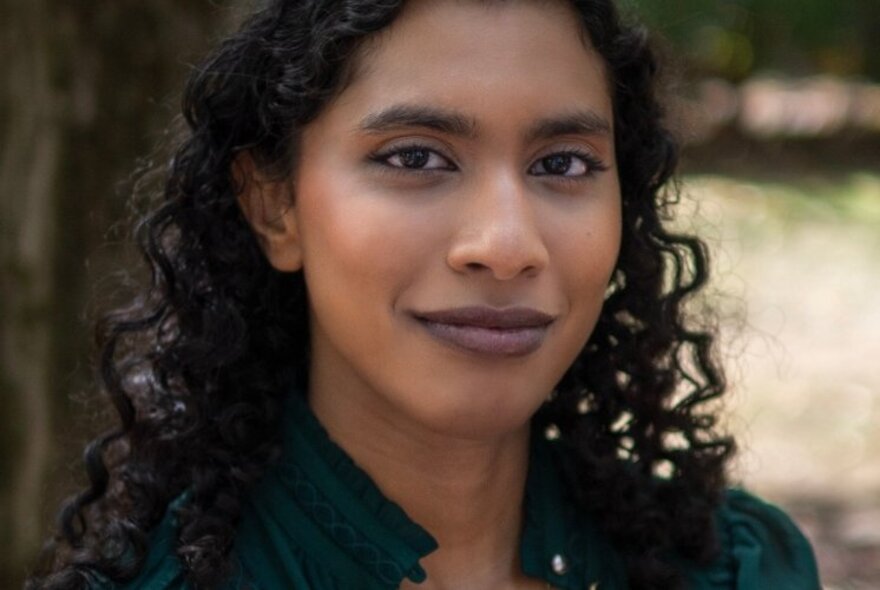 Young woman with long curled black hair and a warm smile, standing outside.