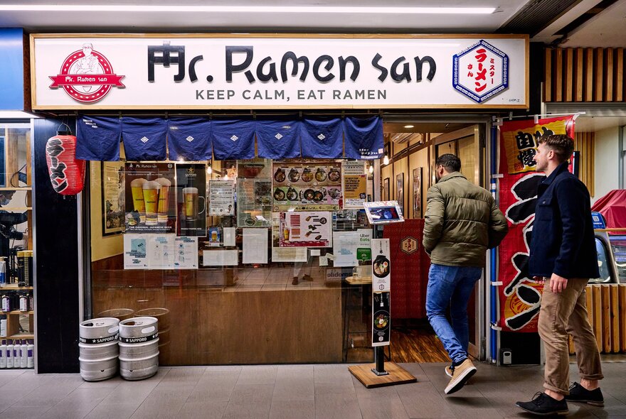 Two friends are walking into a small ramen shop there is a sign above them saying Mr Ramen San.