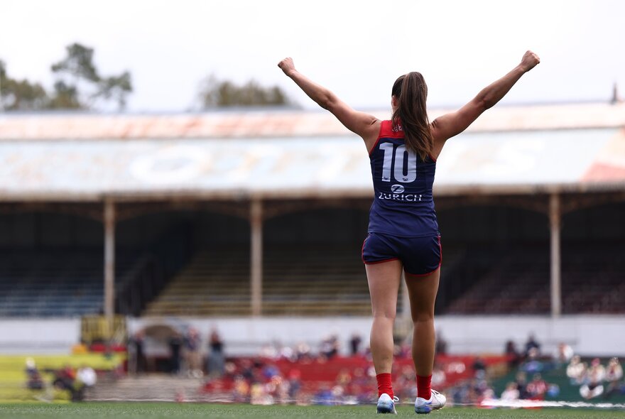A Melbourne Demons AFLW player with her arms in the air in celebration during a game.