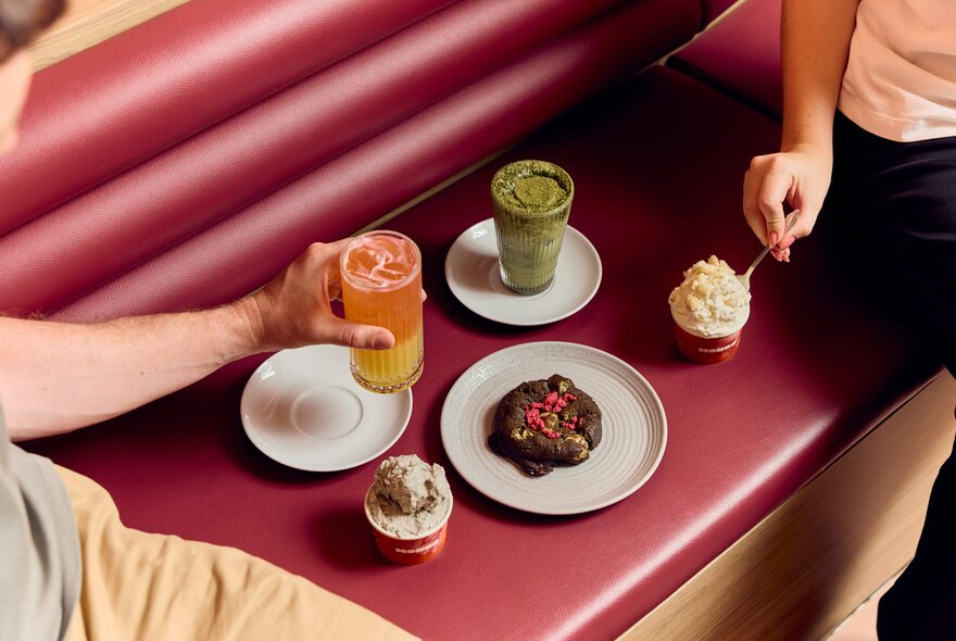 A close-up of a red upholstered bench seat with two people reaching for various desserts and drinks.