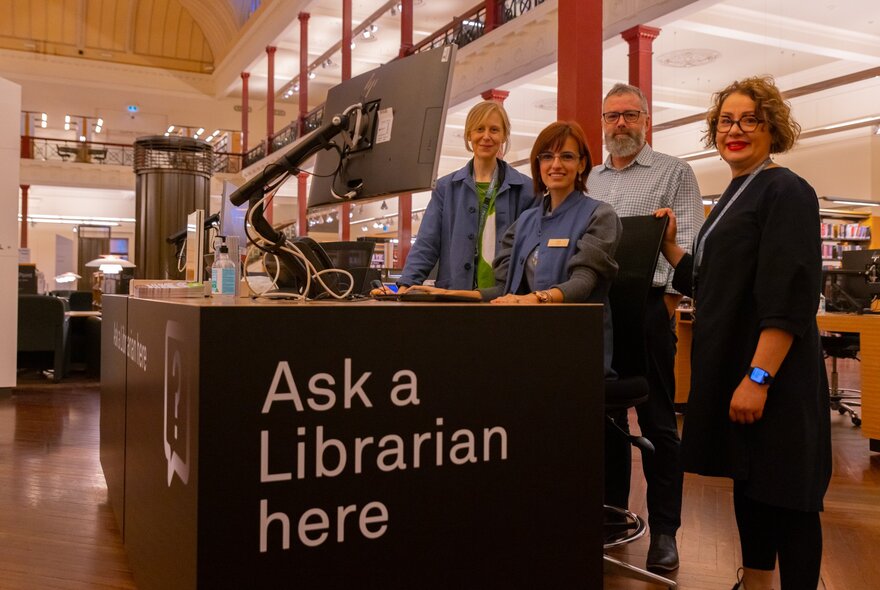 Four librarians standing inside the State Library Victoria, behind a desk that has the words 'Ask A Librarian Here' written on the side of the desk.