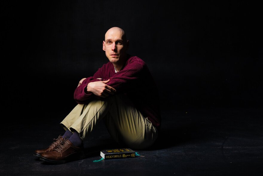 Male actor sitting on the floor of a theatre stage with his arms wrapped around his raised knees, a book on the floor beside him.