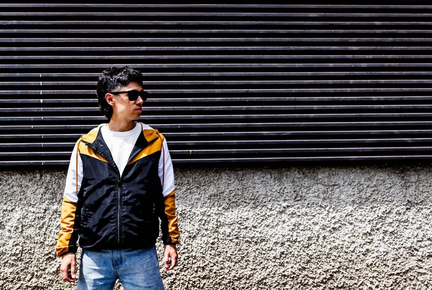 A young person standing in front of a roller door, wearing a 1980s style tracksuit top and dark glasses.