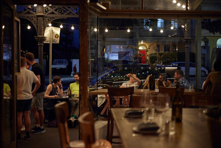 Interior of a small neighbourhood wine bar, a few people standing or sitting around, the view looks out onto the street through large glass windows.