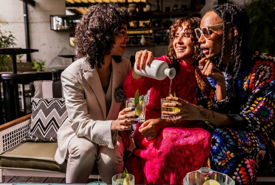 Three women seated on a bench in a bar environment with one woman pouring another a drink.