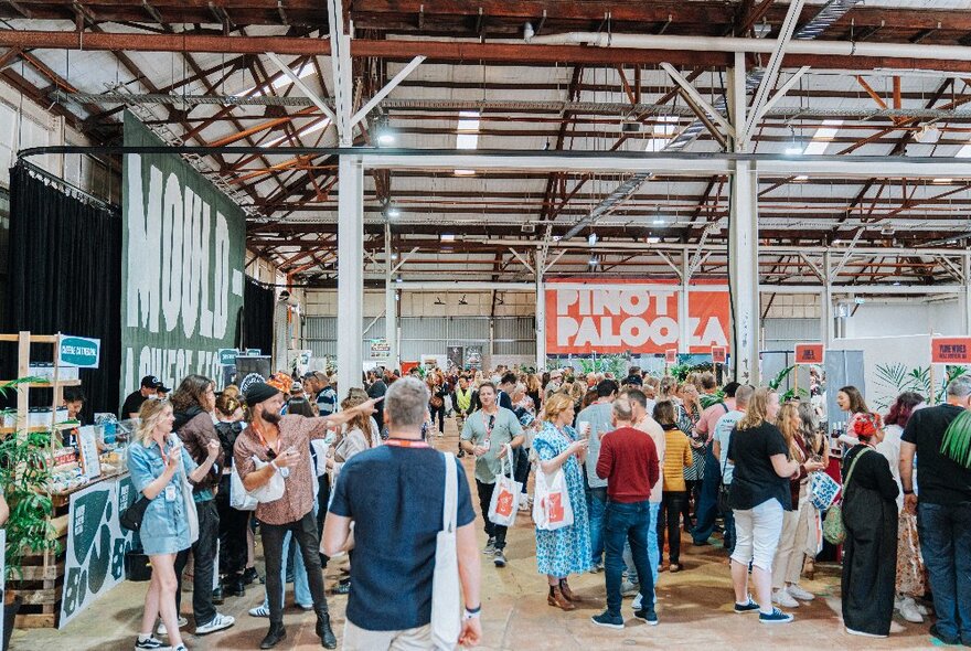 A crowd of people at a food and wine festival inside a large hall, with a sign on the rear wall that reads PINOT PALOOZA.