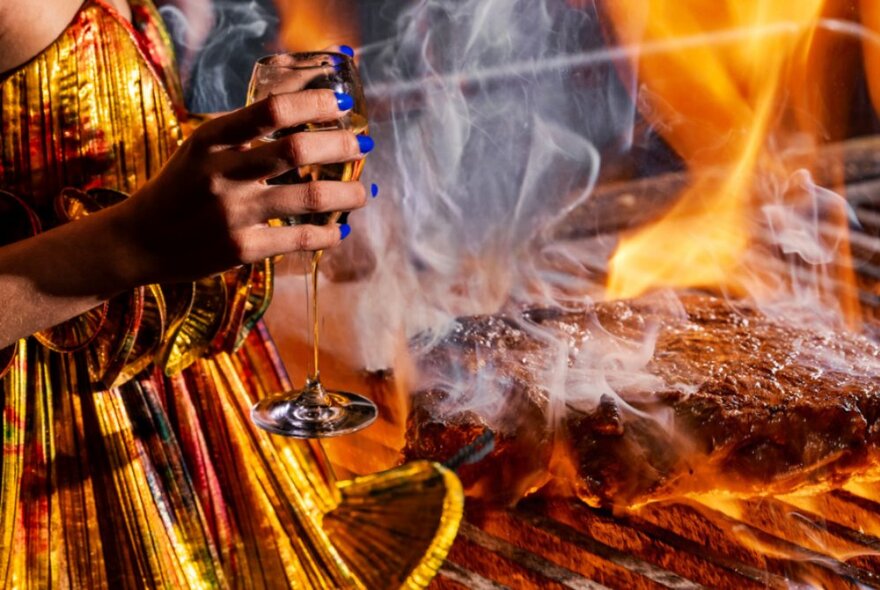 Part of a person wearing a gold dress and holding a glass of sparkling, standing next to a flaming steak on a BBQ.