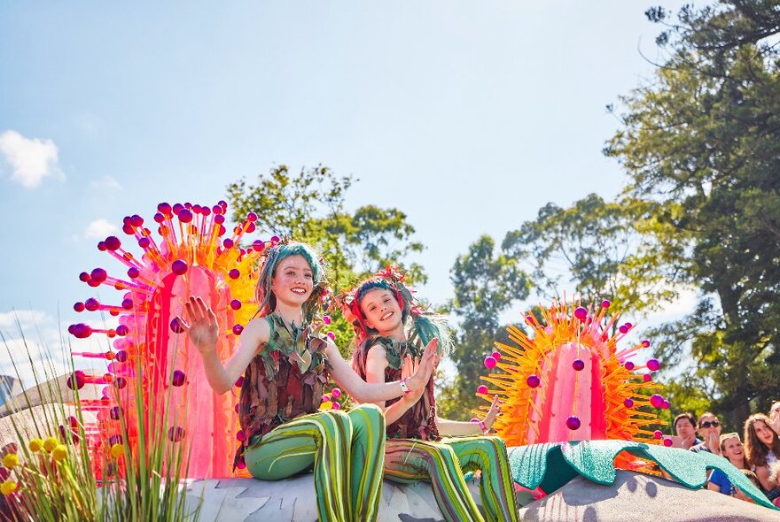 Two young adults dressed as elves, sitting on a float and waving.