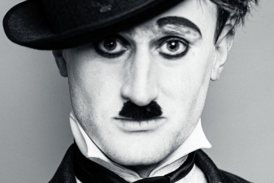 Close up of an actor's face in the character of Charlie Chaplin, with a short cropped moustache, black eyeliner accentuating his eyes and eyebrows, and wearing a black bowler hat.